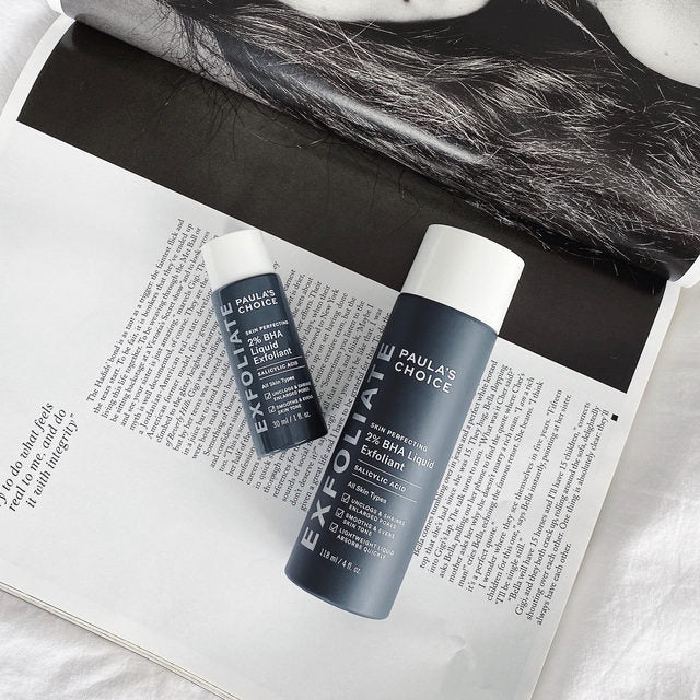 Two sleek bottles of Paulas Choice SKIN PERFECTING BHA Liquid Exfoliant, ideal for blackhead removal, rest on an open magazine with black and white hair imagery. Infused with salicylic acid, this modern product exfoliates dead skin effortlessly.