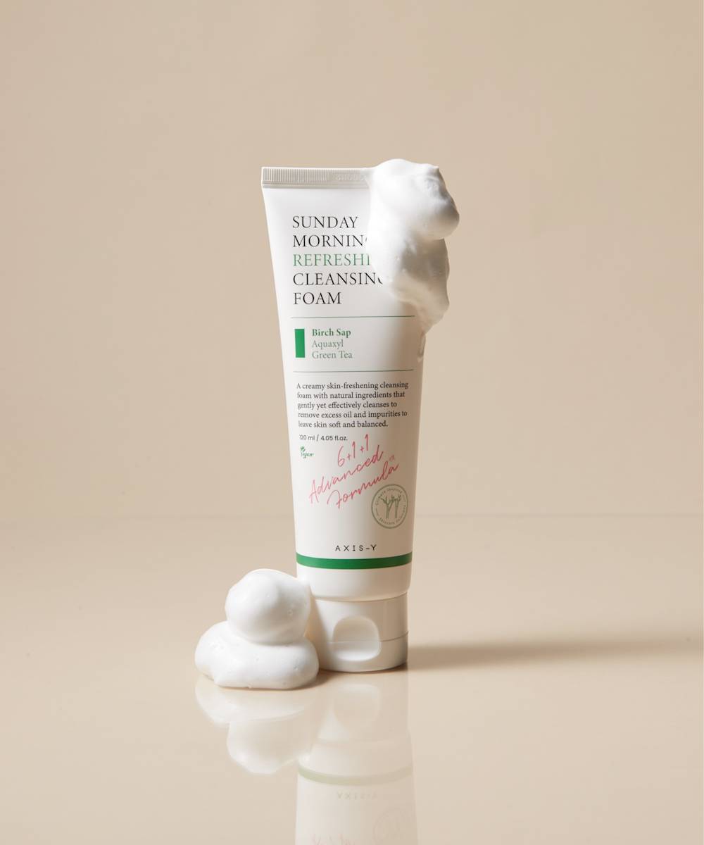 The AXIS-Y Sunday Morning Refreshing Cleansing Foam 120ml is upright on a beige surface, featuring Aquaxyl care technology for sensitive skin. A foam dollop drapes over the top, pooling at the base. The white packaging is embellished with cheerful green and pink text.