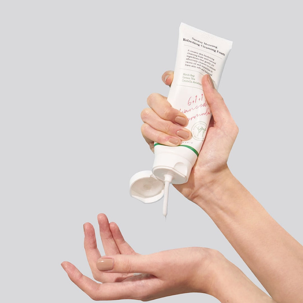 A pair of hands gently squeezes a tube labeled Axis-Y Sunday Morning Refreshing Cleansing Foam 120ml by AXIS-Y, dispensing a small amount perfect for sensitive skin. The white label details its Aquaxyl care technology with text and graphics.