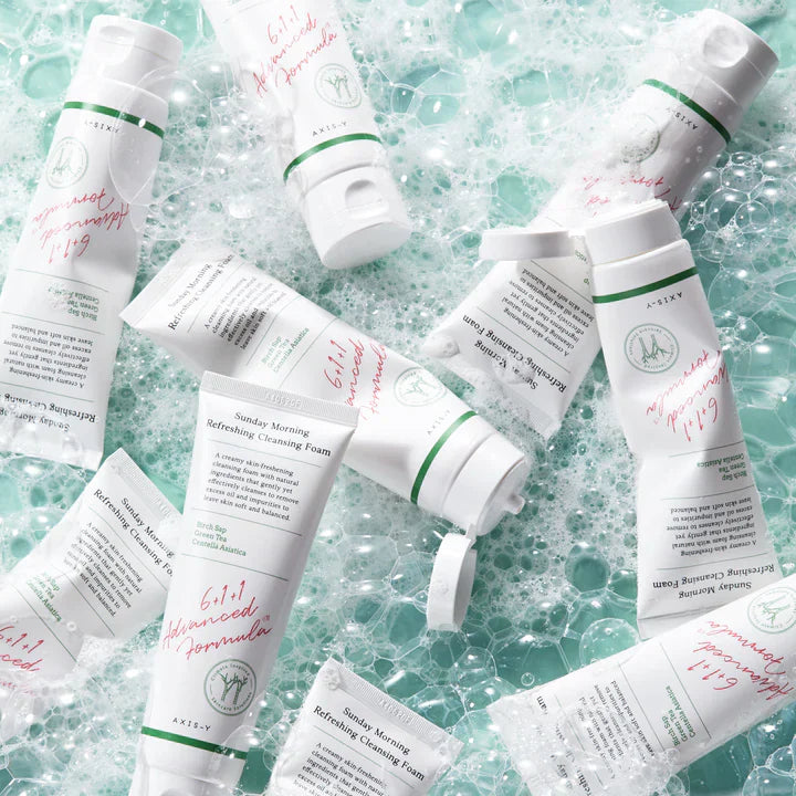 The Axis-Y Sunday Morning Refreshing Cleansing Foam 120ml sits on a bubbly, water-like surface. Tailored for sensitive skin, the white tube features green and pink text with a botanical illustration and utilizes Aquaxyl care technology for gentleness.