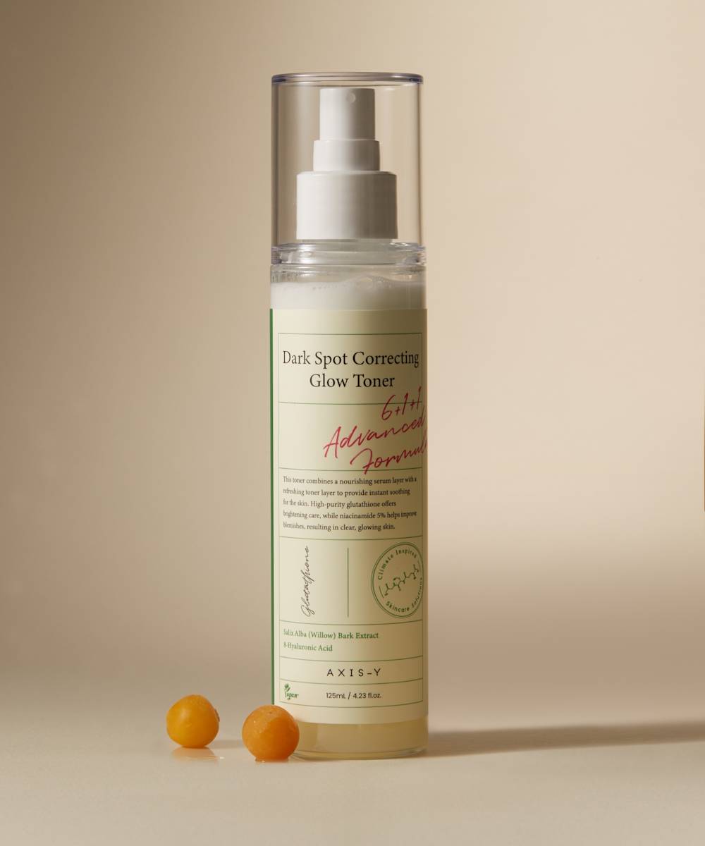 The Axis-Y Dark Spot Correcting Glow Toner by AXIS-Y comes in a bottle with white and red text on a light background, featuring a pump dispenser and round cap. It includes botanical drawings and essential skincare info, alongside two small orange spheres.