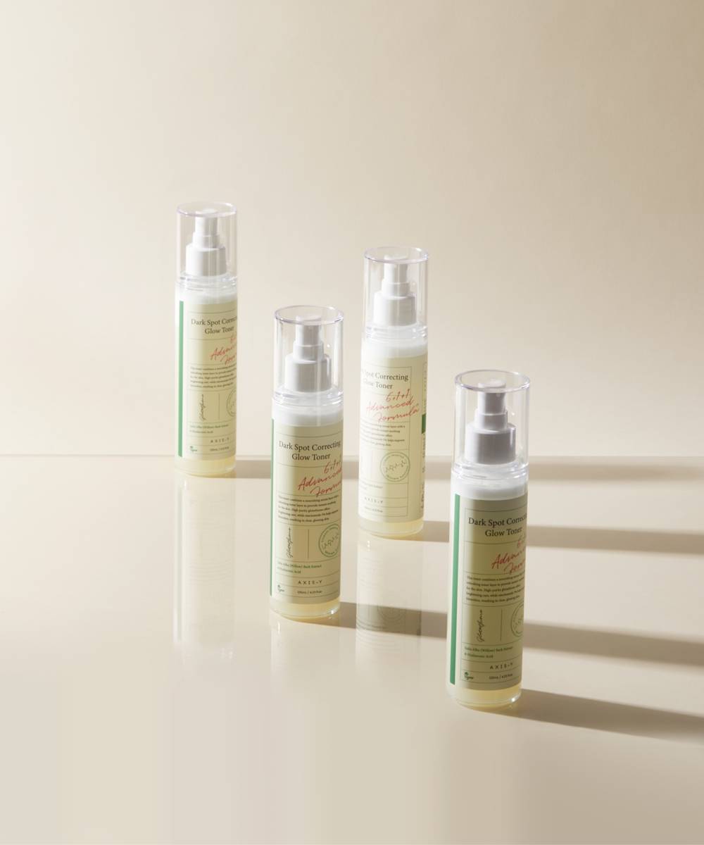 Four bottles of Axis-Y Dark Spot Correcting Glow Toner, featuring pumps and green labels with text and botanical graphics, are arranged on a reflective surface against a neutral background, highlighting their focus on natural ingredients for an even-toned complexion.