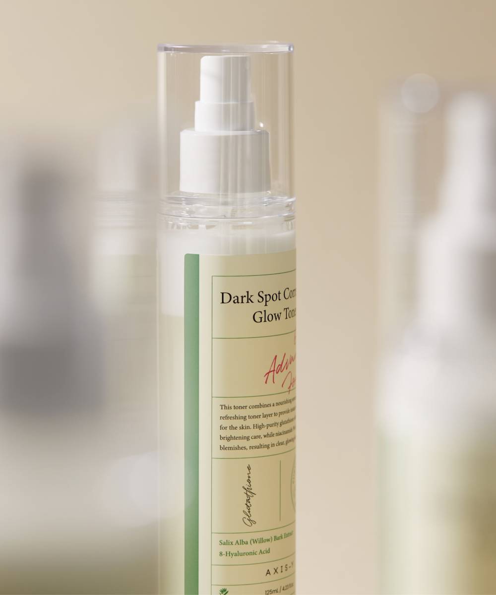 The Axis-Y Dark Spot Correcting Glow Toner 125ml by AXIS - Y is showcased against a blurred beige backdrop. This brightening toner in a clear bottle with a white pump features light-colored liquid and highlights ingredients like Salix Alba and Hyaluronic Acid for enhanced radiance.