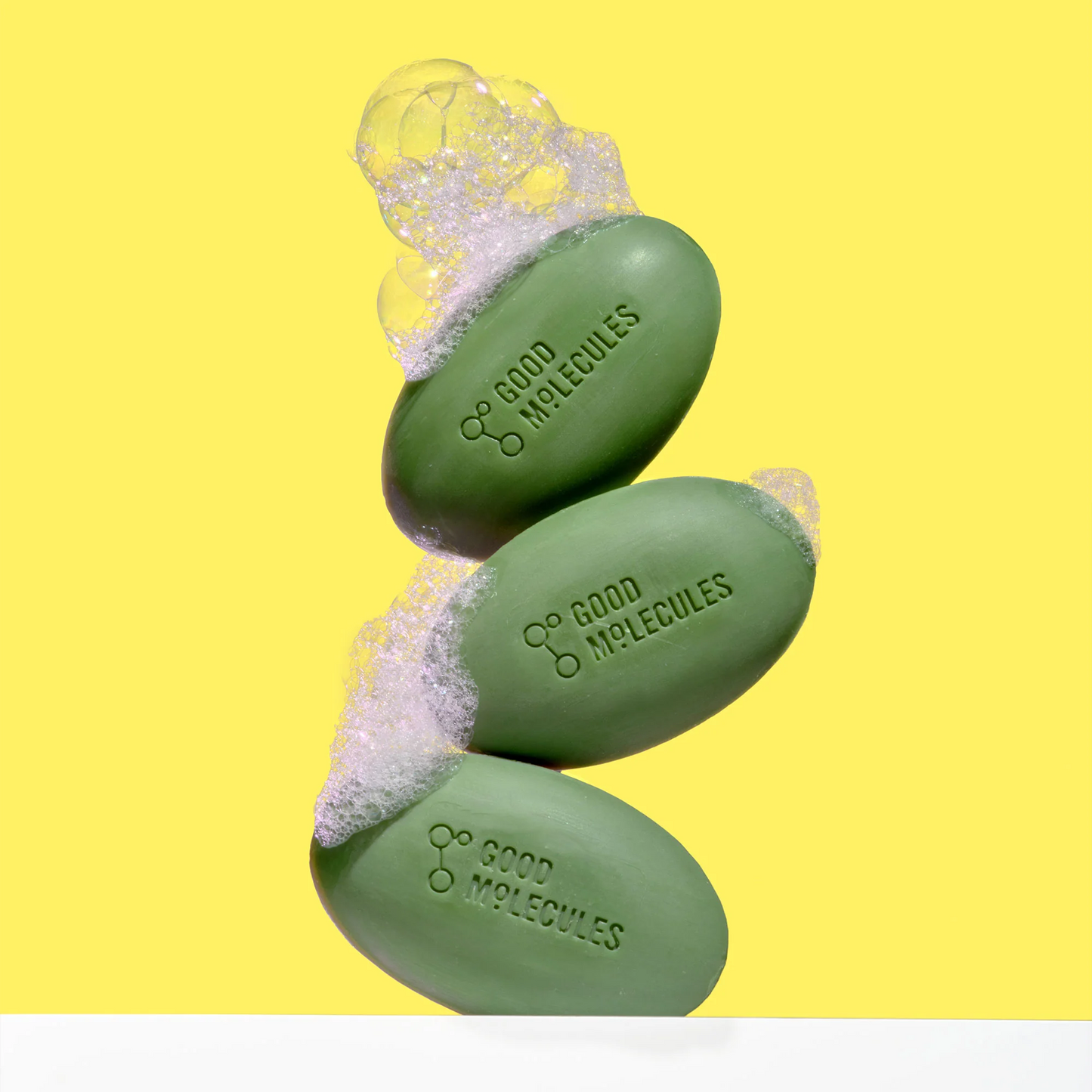 Three green bars of Good Molecules Brightening & Dark Spots Bar with Kojic Acid - 85g, featuring their logo, are stacked against a vibrant yellow background. Bubbles atop the bars enhance its fresh appeal, ideal for combating dark spots and skin discoloration for a brightening effect.