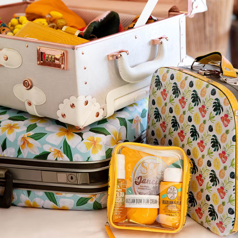 Sol de Janeiro Brazilian Bum Bum Jet Set features colorful suitcases, one with a tropical floral design, and a floral-patterned toiletries bag. A clear pouch holds travel minis, including the iconic Sol de Janeiro Brazilian Bum Bum Cream in vibrant yellow packaging with the Cheirosa 62 scent.
