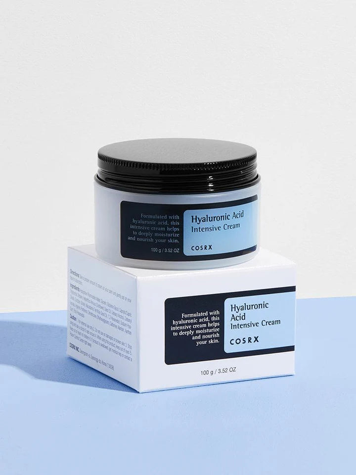 COSRX Hyaluronic Acid Intensive Cream jar placed on its box against a soft blue background. A rich, hydrating formula for deep moisture and skin repair.