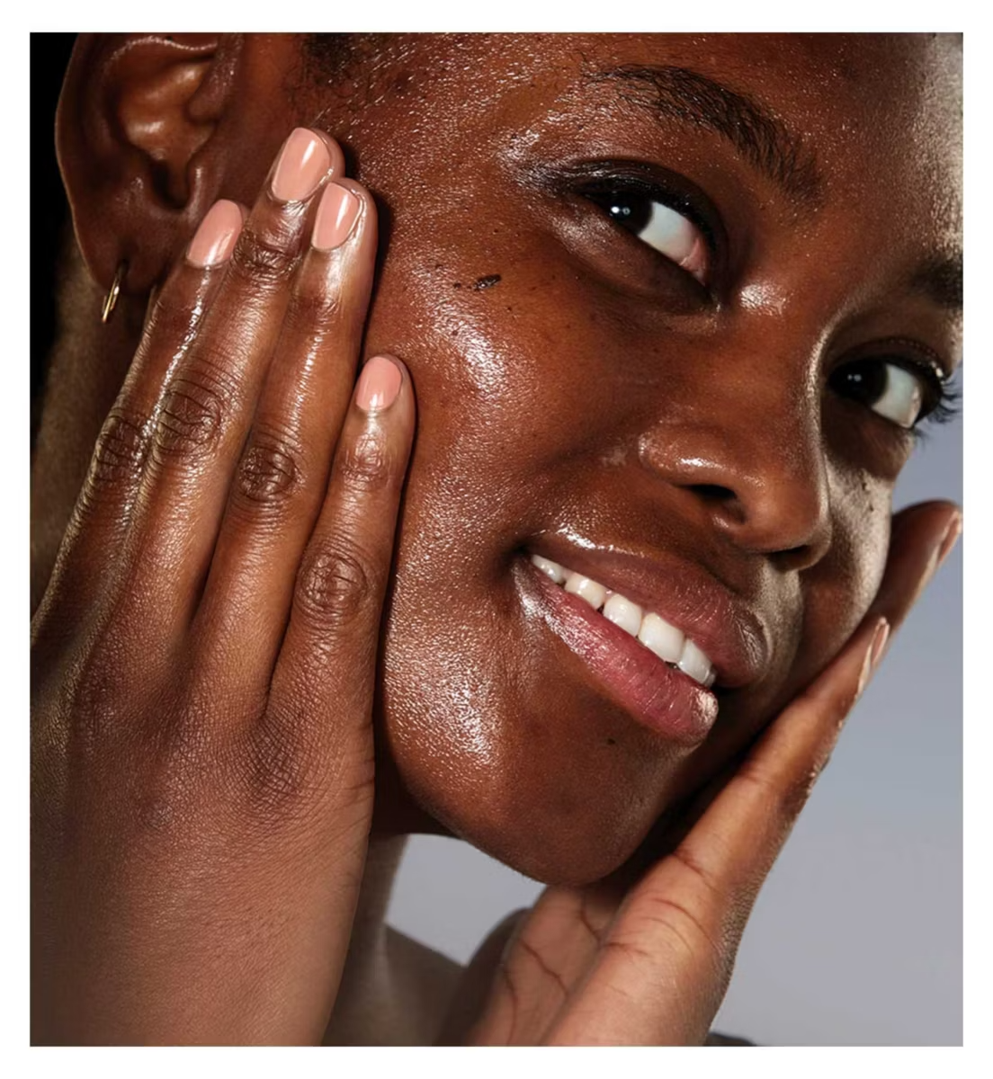 A close-up of a smiling person with glowing, dewy skin touches their cheeks, radiating the nourishing effects of ESTEE LAUDER’s Advanced Night Cleansing Balm. Their neatly manicured nails are visible as they gaze slightly upward.