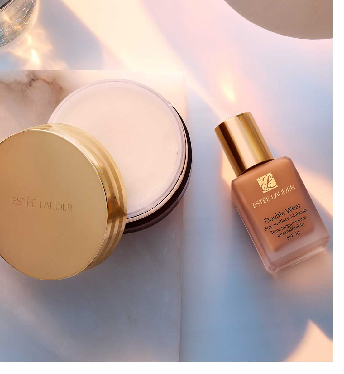 Two ESTEE LAUDER beauty products on a marble surface: Advanced Night Cleansing Balm in a round jar with a gold lid and Double Wear Stay-in-Place Makeup foundation, both highlighted by soft lighting that enhances their luxurious packaging.