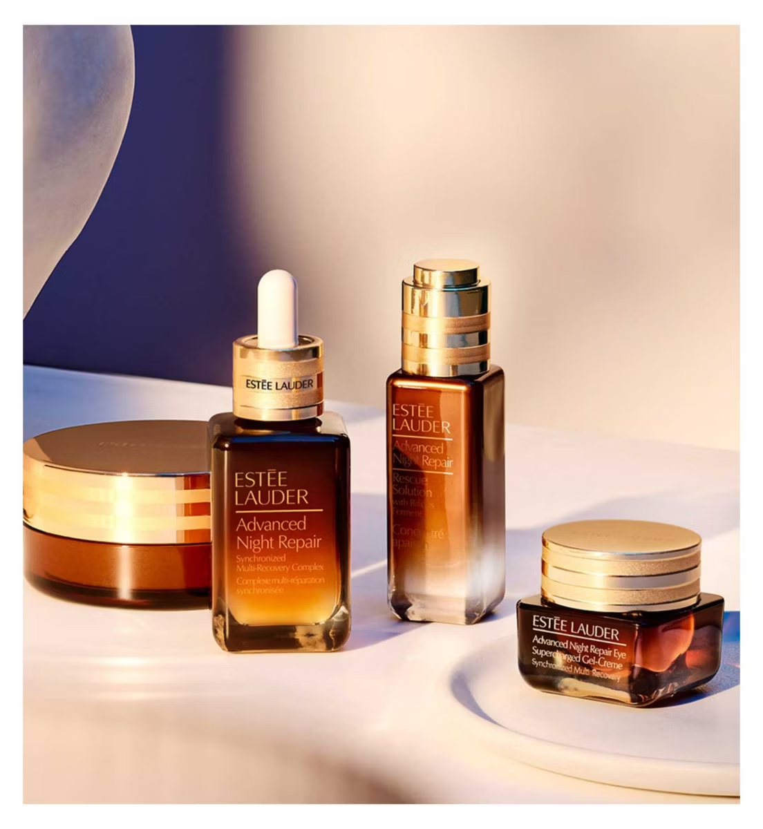 Four ESTEE LAUDER skincare products, including Advanced Night Repair and the Estée Lauder Advanced Night Cleansing Balm, are arranged on a light surface in warm, soft lighting, with gold-capped bottles and jars highlighted.