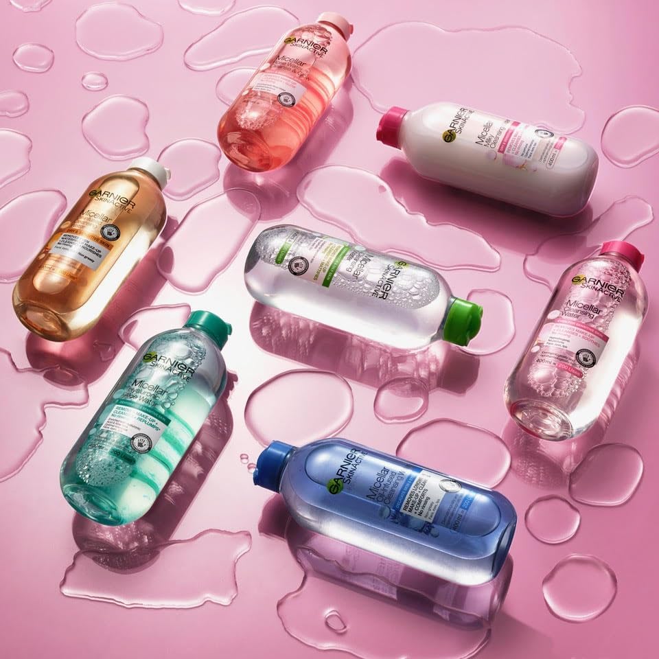 Seven bottles of Garniers Micellar Cleansing Water, an ideal makeup remover and facial cleanser for combination and oily skin, are arranged on a wet pink surface. Each bottle has a differently colored cap—green, red, pink, and blue—adorned with sparkling water droplets.