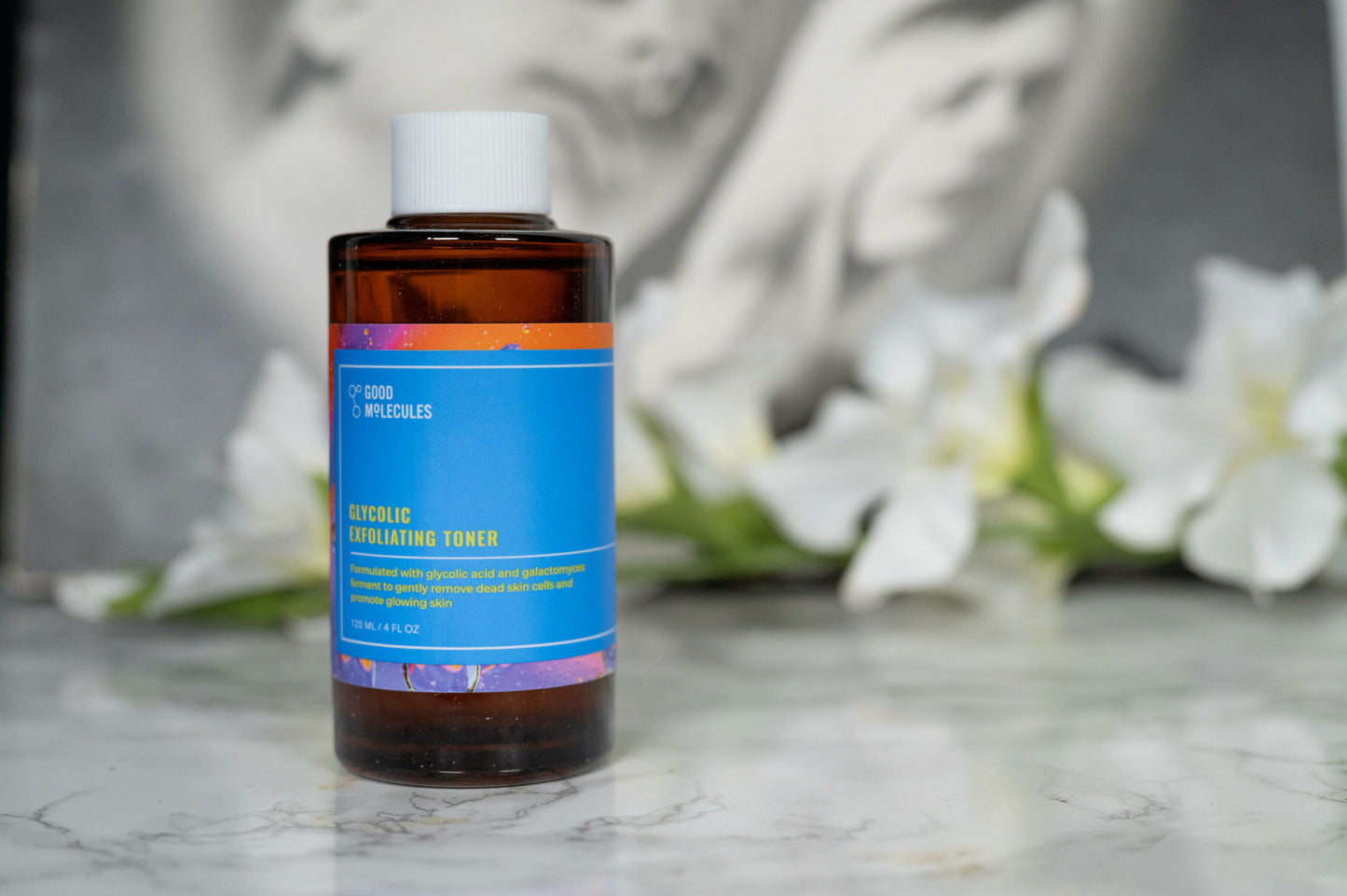 A brown Good Molecules Glycolic Exfoliating Toner bottle with a blue label rests elegantly on a marble surface, with softly blurred white flowers in the background, enhancing the serene vibe of this skincare staple.