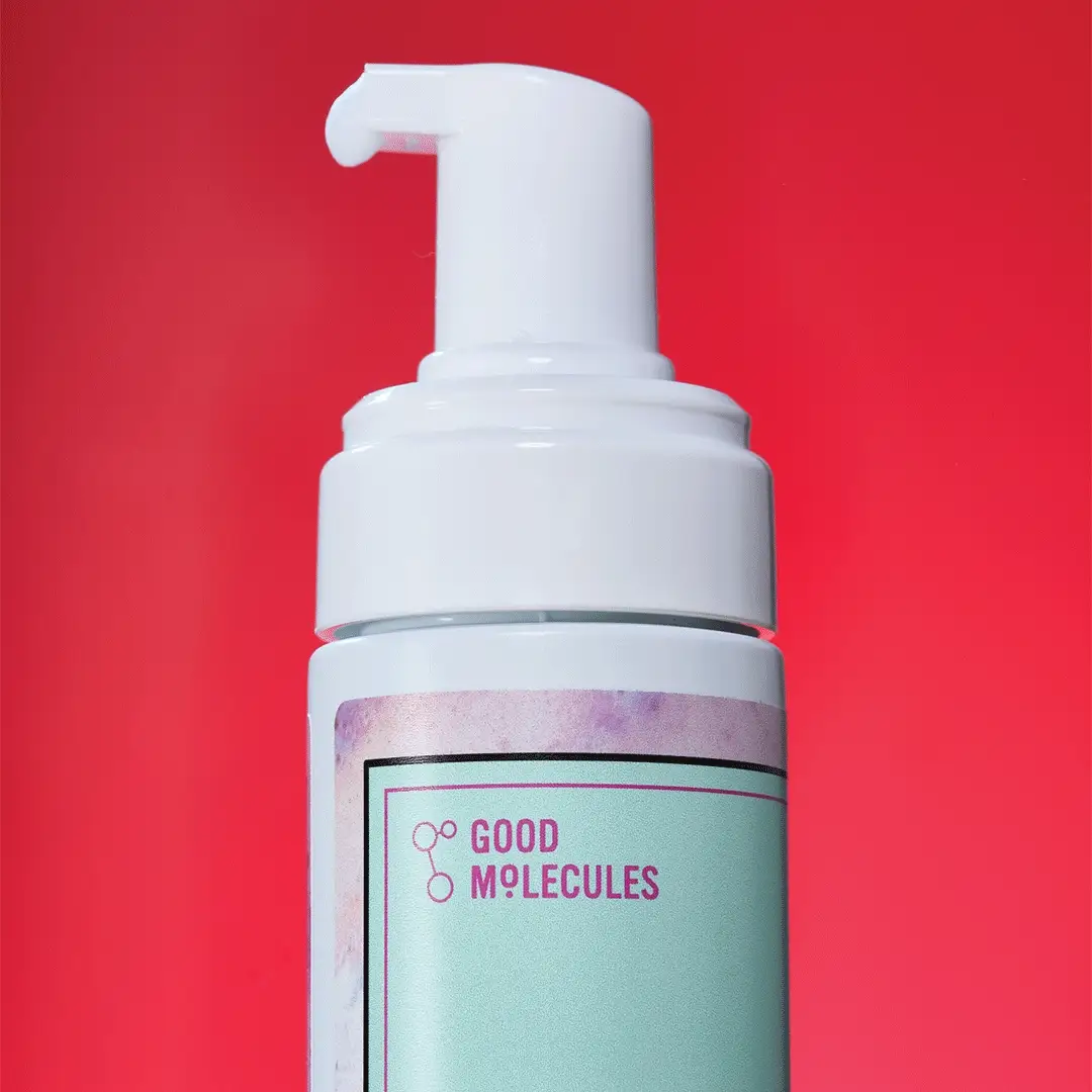 A bottle of Good Molecules Acne Foaming Cleanser with Salicylic Acid BHA, featuring a white pump, stands against a vibrant red backdrop. The label displays the brand name in purple text with molecular graphics.