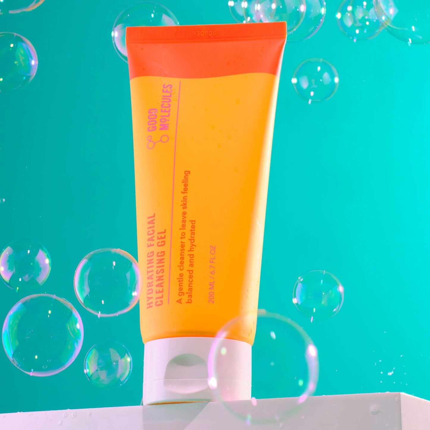 A vibrant orange tube of Good Molecules Hydrating Facial Cleansing Gel, 200ml, stands upright on a white surface with floating soap bubbles around it. The turquoise blue background enhances the lively hues of the packaging.