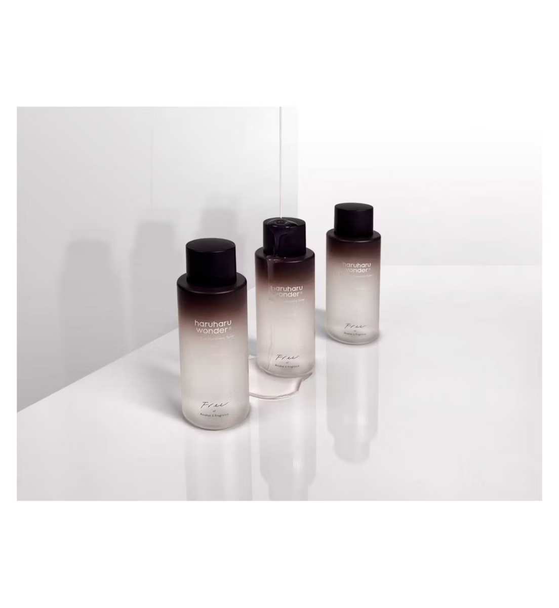 Three bottles of Haruharu WONDER Black Rice Hyaluronic Toner sit on a glossy white surface. Their minimalist design features a clear-to-dark gradient topped with black caps, casting soft shadows that reflect their elegance. Each bottle is 150ml and fragrance-free.