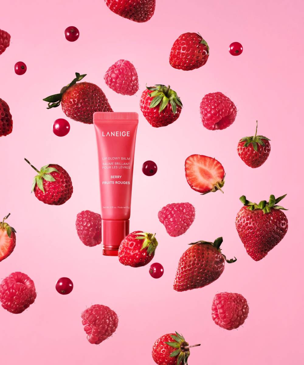 A pink tube of Laneige Lip Glowy Balm - Berry 10g, enriched with shea and murumuru butters, is surrounded by floating strawberries, raspberries, and red currants on a pink background, creating a vibrant fruity aesthetic.