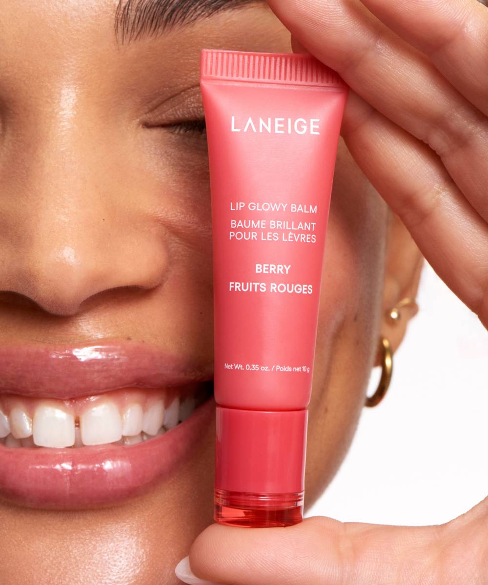 A person smiles, holding a Laneige Lip Glowy Balm - Berry 10g infused with shea and murumuru butters. The pink tube with white text complements their partly visible face, highlighting luscious lips and one eye against a pristine white background.