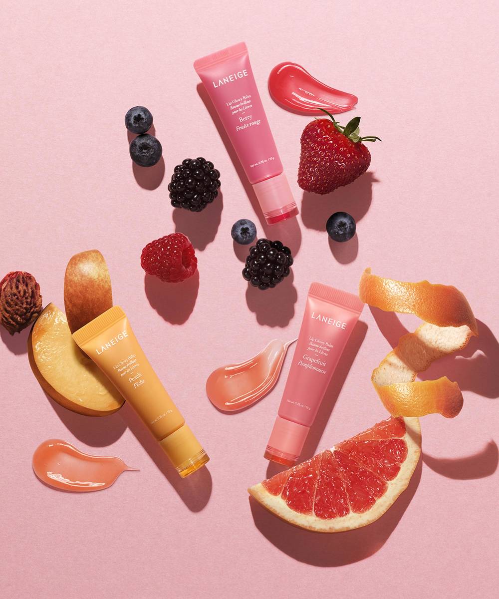 Three tubes of Laneige Lip Glowy Balm - Berry 10g are displayed among berries and fruit slices on a pink background. Infused with shea and murumuru butters, they promise hydration and a hint of color.