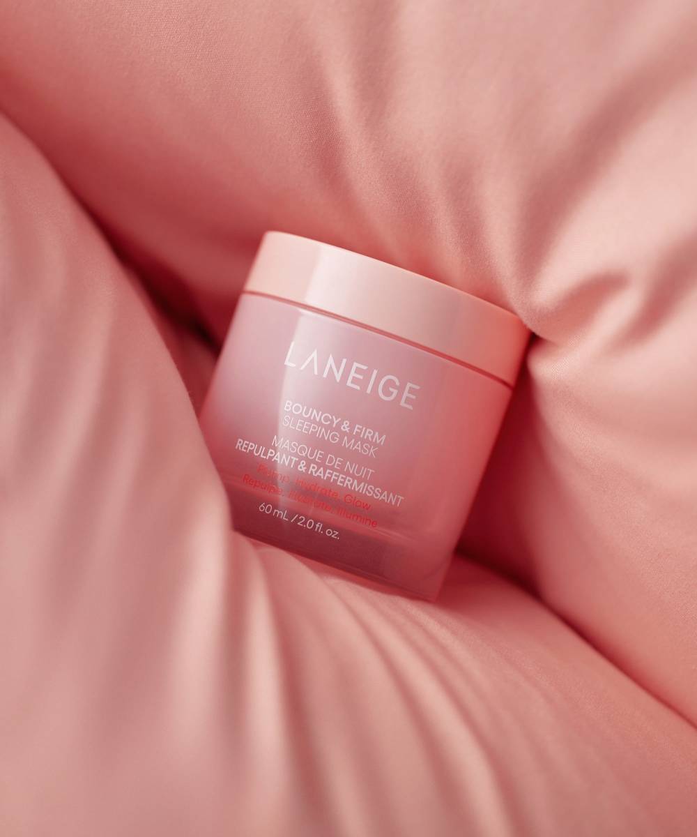 A pink jar labeled Laneige Bouncy & Firm Sleeping Mask rests on soft pink fabric. This 60 ml container exudes luxury and softness, focusing on skincare and relaxation for enhanced skin radiance while you sleep.