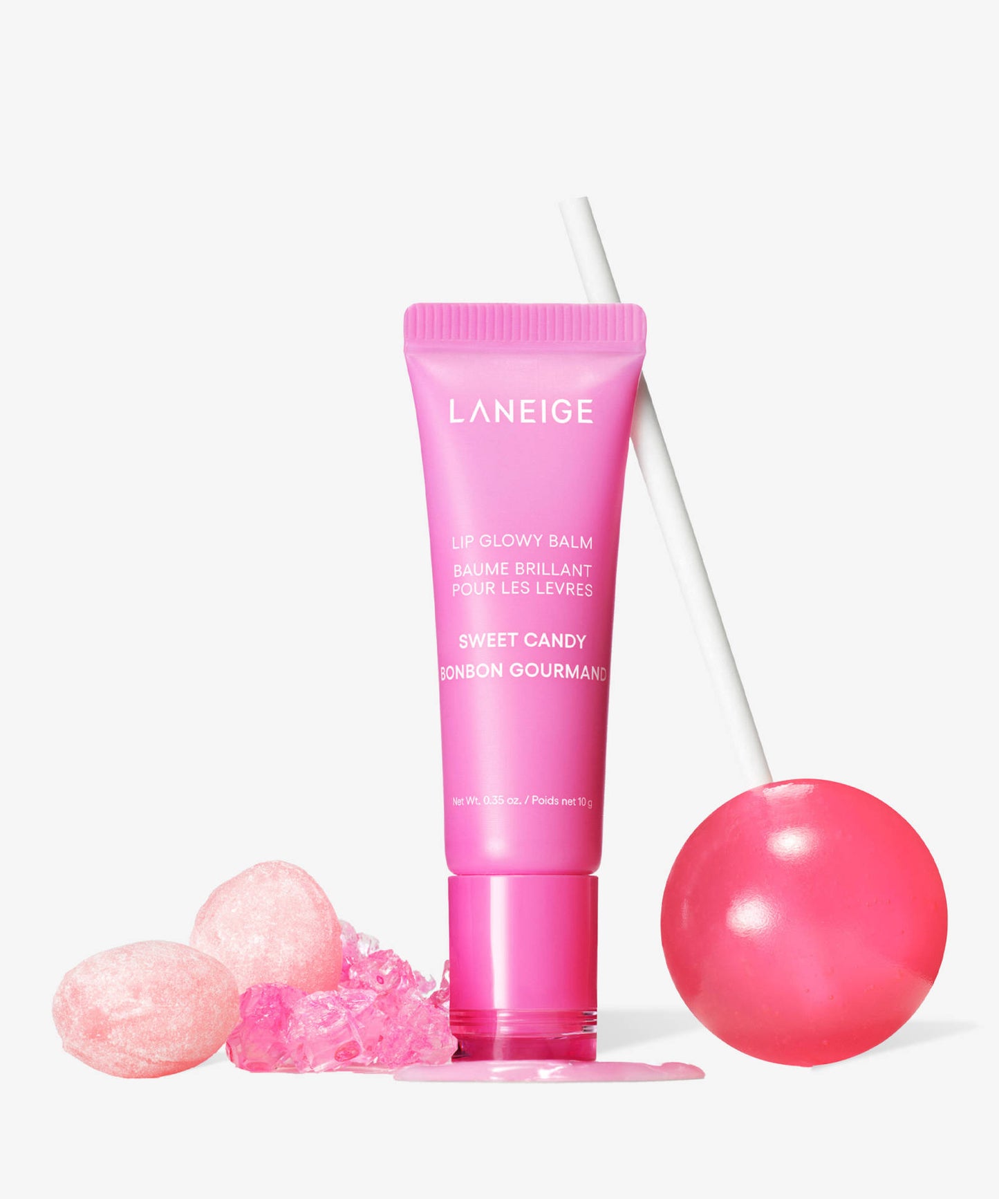 A pink tube of Laneige Lip Glowy Balm - Sweet Candy 10g stands upright, featuring shea and murumuru butters for a glass skin glow. Beside it are a shiny pink lollipop and two small pink candies, complementing the sweet theme against a white background.