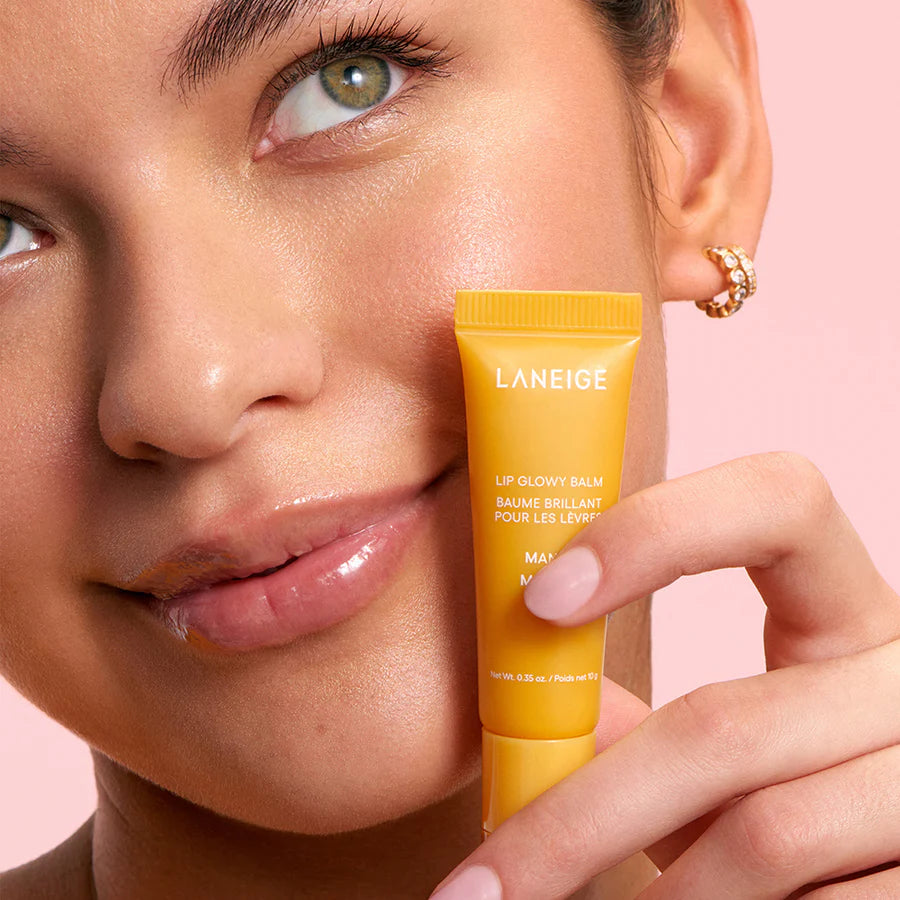 A person showcases the hydrating Laneige Lip Glowy Balm - Mango 10g against a pink backdrop while wearing gold hoop earrings. Their smooth, glowing skin mirrors the glossy effect of the LANEIGE product they hold, highlighting its moisturizing power.