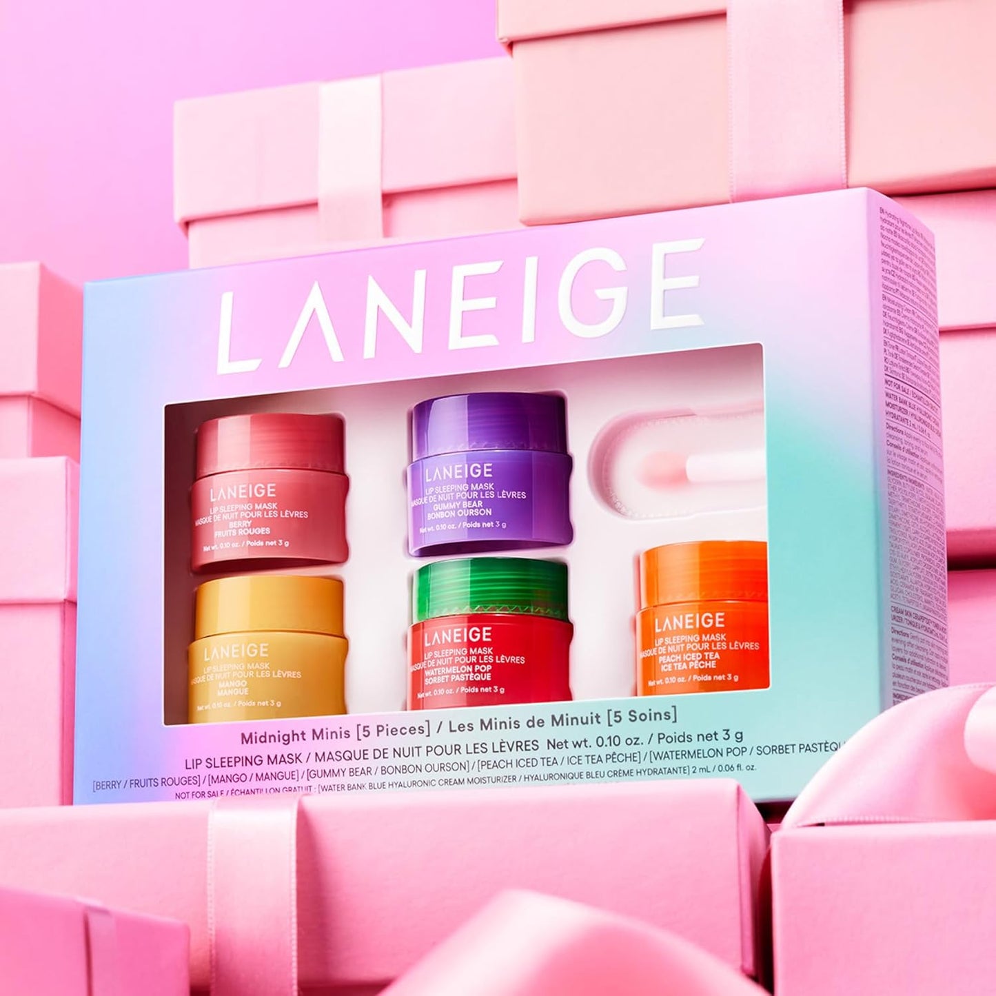 Laneige Midnight Minis Lip Sleeping Mask Set displayed among pink gift boxes. The packaging features a gradient pastel design with the 'LANEIGE' logo, a clear window showcasing five colorful mini jars in pink, purple, yellow, green, and orange, along with an applicator. The set includes flavors such as Berry, Mango, Gummy Bear, Peach Iced Tea, and Watermelon Pop.