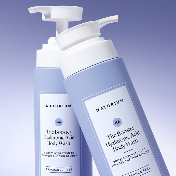 Two 500ml light purple bottles of Naturium The Booster Hyaluronic Acid Body Wash, with white pump dispensers and hydration and fragrance-free highlights, are set against a soft blue background.