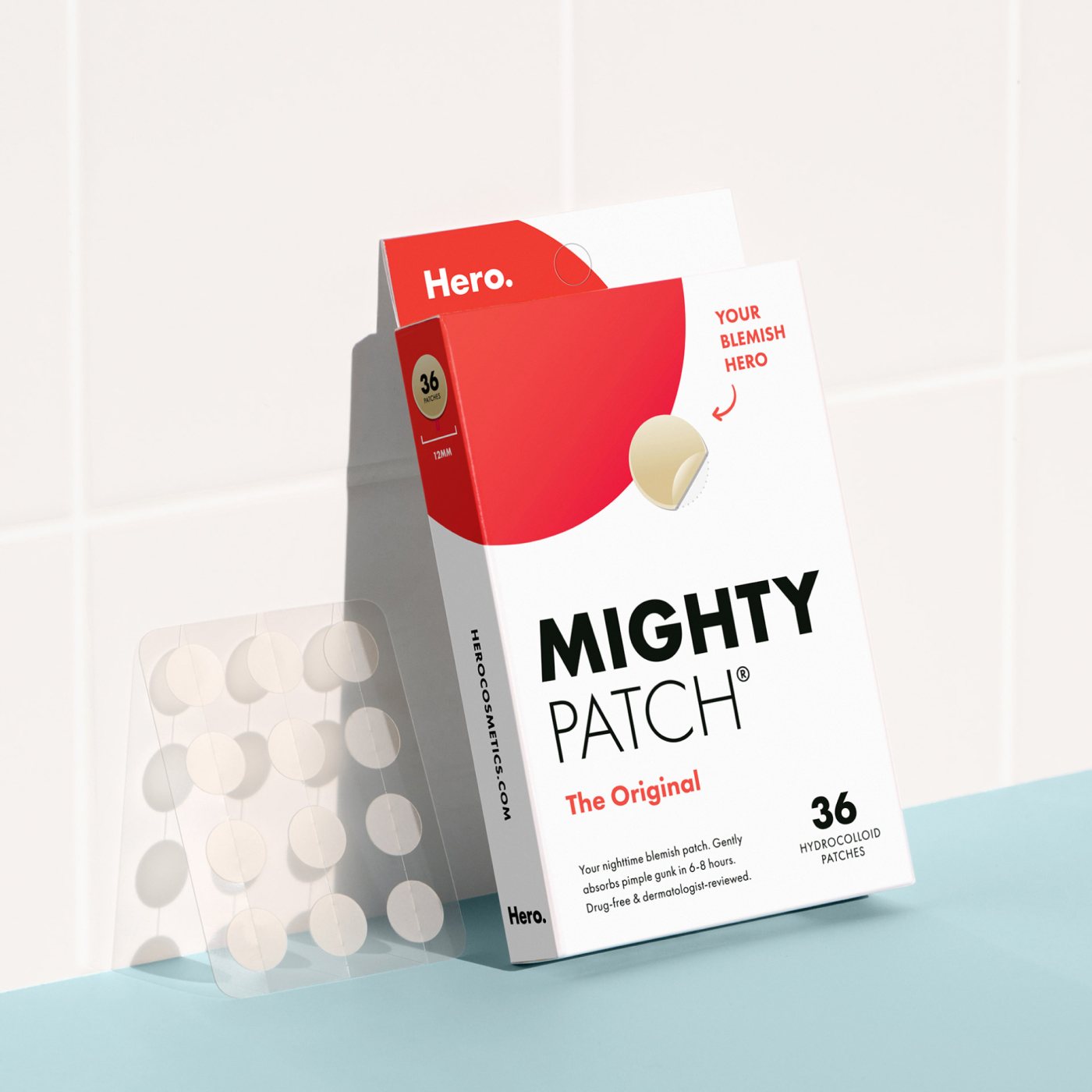 A box of Hero Cosmetics Mighty Patch® The Original contains 36 hydrocolloid acne patches. The packaging is red and white, and a sheet of transparent, circular patches is shown next to the box on a tiled surface.