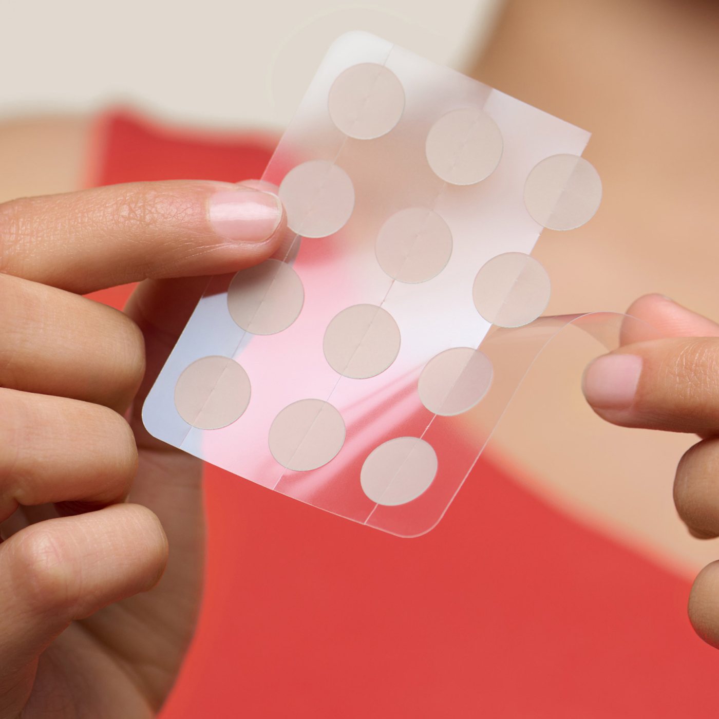 Dressed in a red shirt, a person carefully peels a Hero Cosmetics Mighty Patch® The Original off its clear plastic backing. The round, transparent adhesive patches subtly target blemishes, with the neutral background emphasizing their effectiveness and discretion.