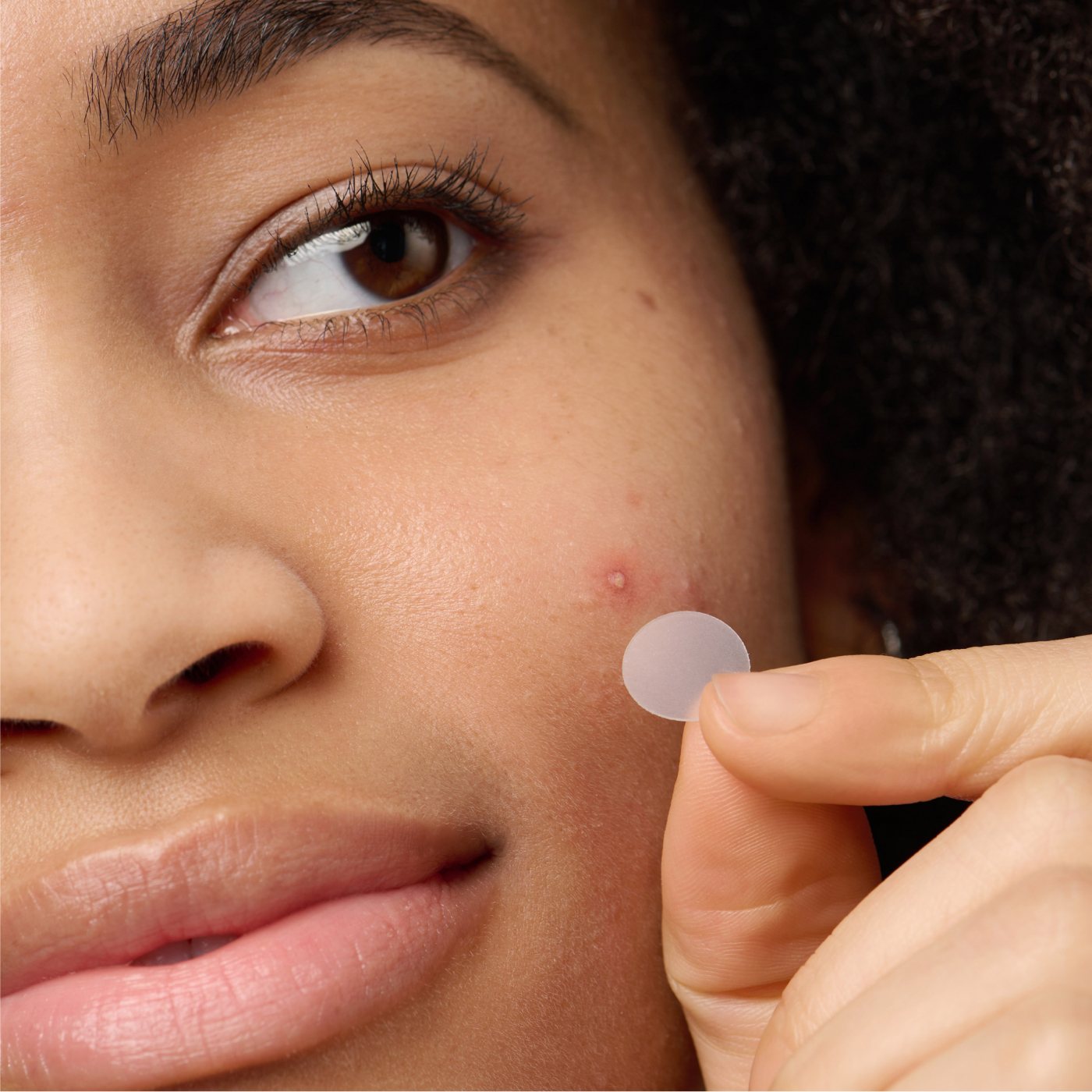 Close-up of a person with curly hair and a calm expression applying the Hero Cosmetics Mighty Patch® The Original to a pimple on their cheek, highlighting an effective skincare routine with Heros 36 clear hydrocolloid patches.