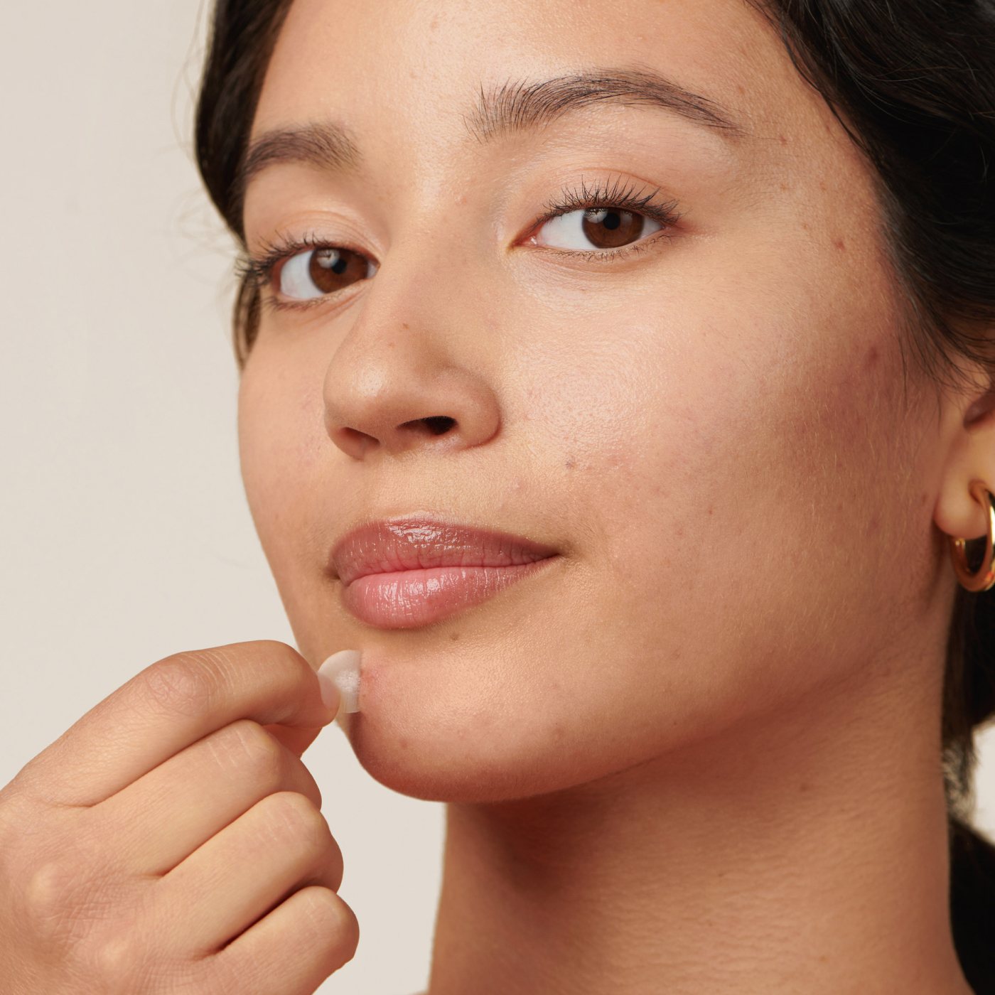 A person with earrings uses a Hero Cosmetics Mighty Patch® on their chin. They have a natural complexion with visible skin texture, looking neutrally at the camera against a plain background.