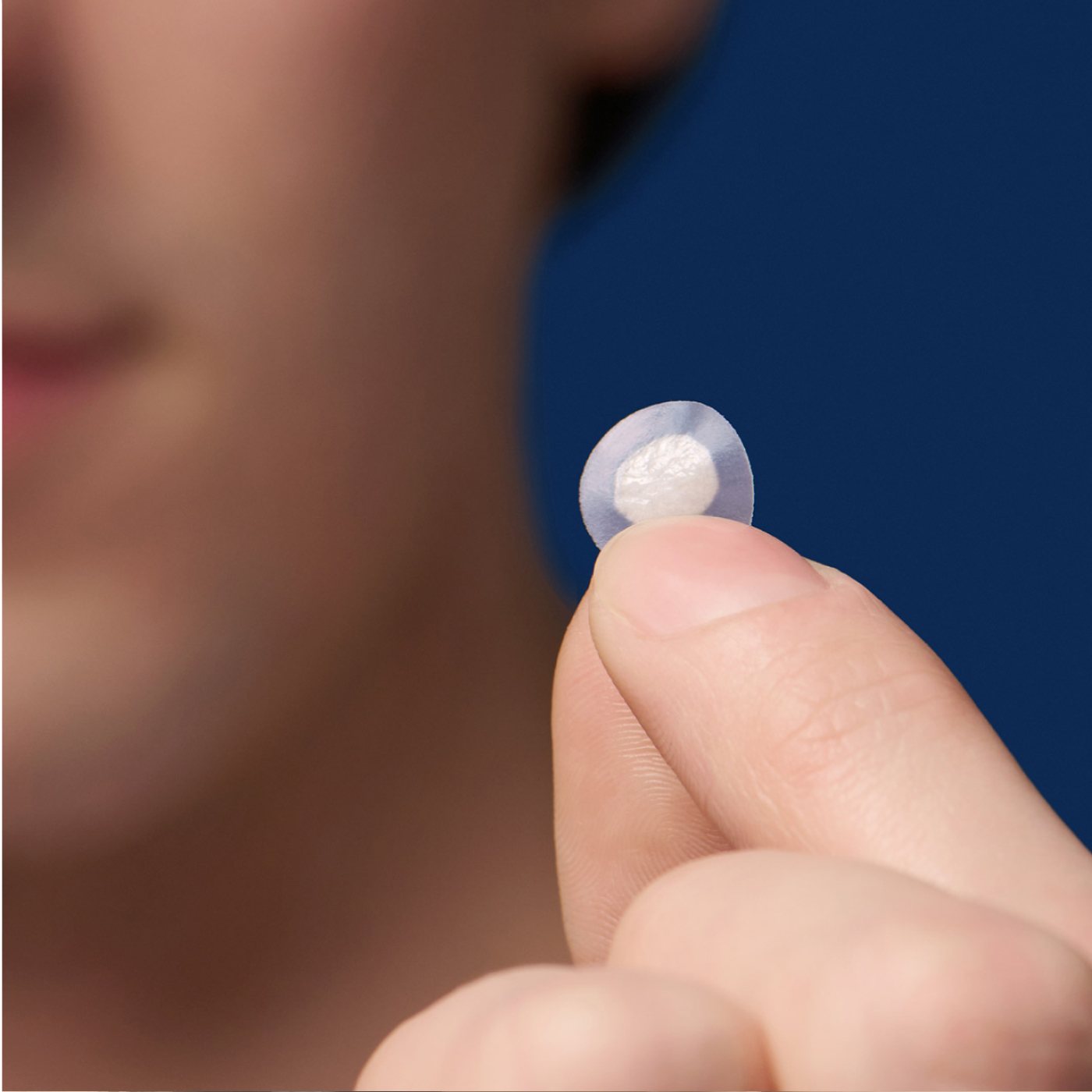 A person holds a small, round, transparent hydrocolloid patch from Hero Cosmetics Mighty Patch® The Original - 36 Patches between their thumb and forefinger against a blue background.