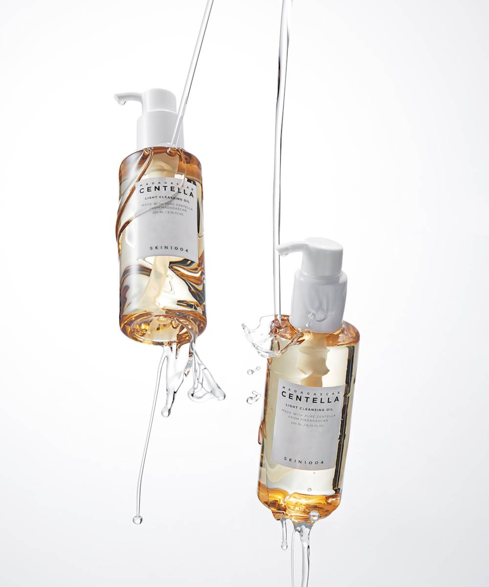 Two bottles of SKIN1004 Madagascar Centella Light Cleansing Oil suspended in mid-air with water splashing around them against a white background.