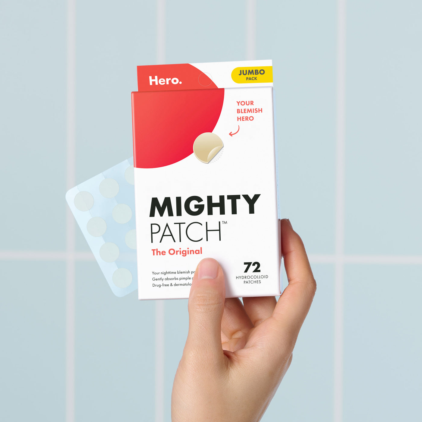 A hand holds a box of Hero Cosmetics Mighty Patch® The Original, labeled 36 Hydrocolloid Patches. This acne patch set comes in a white box with a red top and features circular samples. The words Your Blemish Hero appear on the front against a blue tiled background.