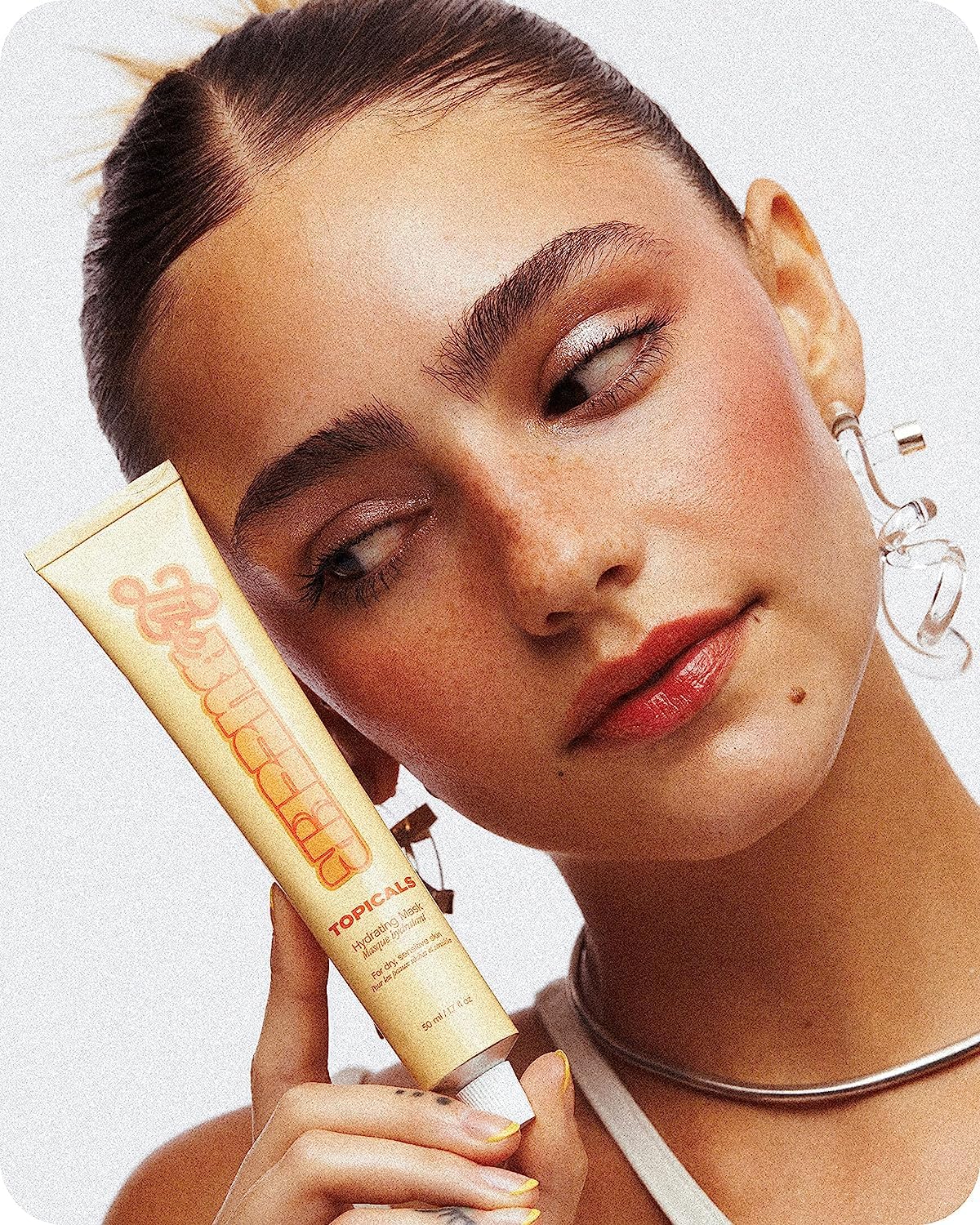 A young woman with groomed eyebrows, dewy skin, and a sleek hairstyle holding a tube of Topicals Like Butter Hydrating Moisturizer close to her face. She has a confident expression, glossy lips, and is wearing clear sculptural earrings.