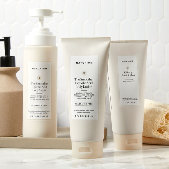 Three Naturium skincare products, including The Smoother Glycolic Acid Body Lotion (234ml), Glycolic Acid Body Wash, and KP Body Scrub & Mask, are displayed on a bathroom counter with a white tile background.