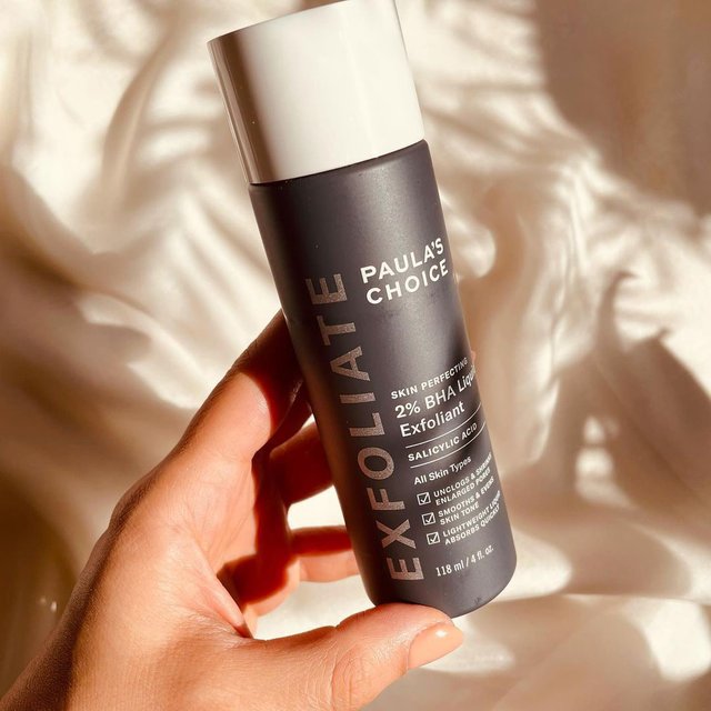 A hand holds a bottle of PAULAS CHOICE SKIN PERFECTING BHA Liquid Exfoliant, renowned for its salicylic acid effectiveness in removing dead skin, against a softly lit fabric background. The dark bottle has white and gray text highlighting its blackhead removal benefits.