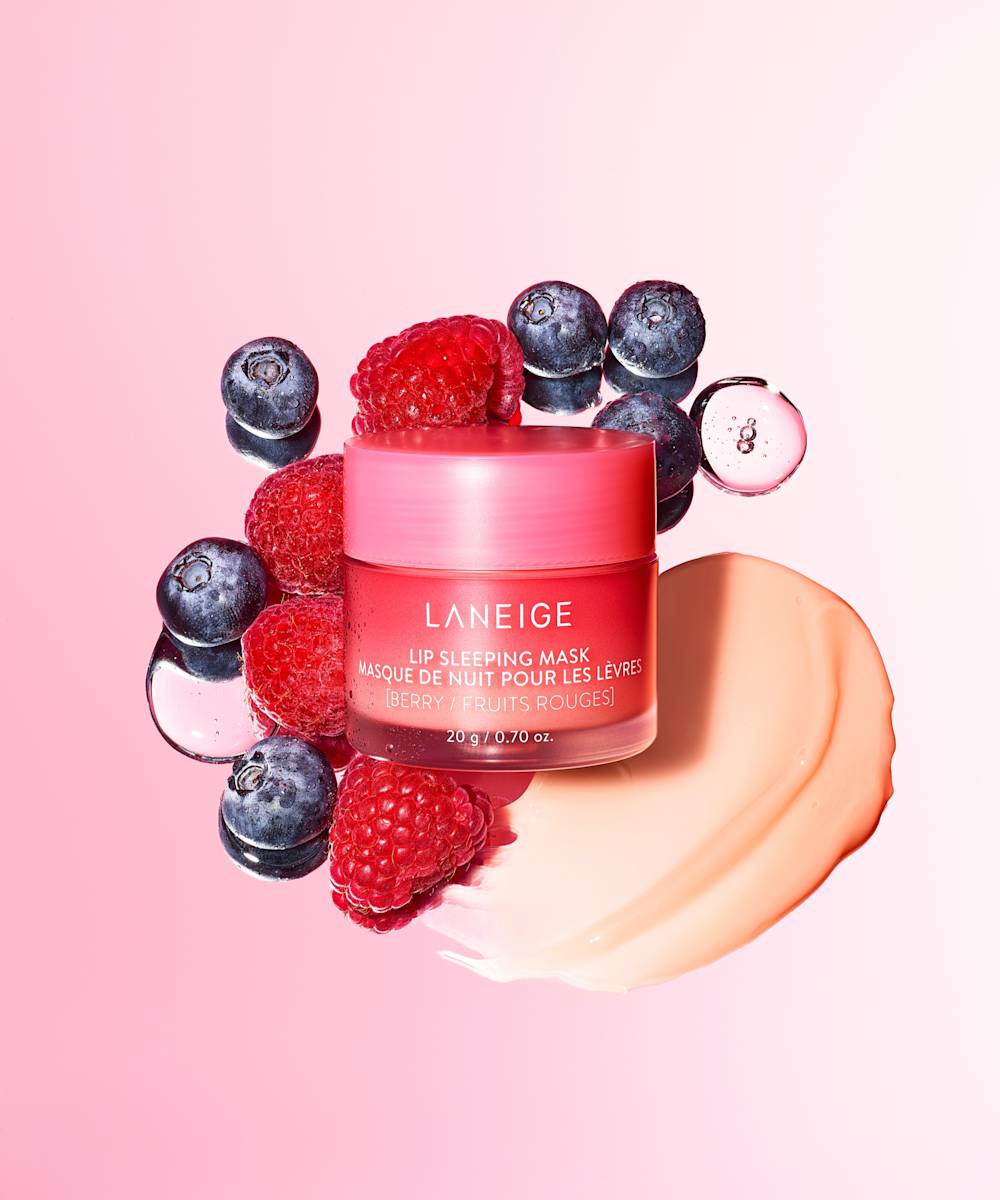 A pink jar of LANEIGEs Laneige Lip Sleeping Mask - BERRY 20g, rich in vitamin C, is surrounded by fresh blueberries and raspberries on a pink background, with a dollop of the creamy product smeared beside it to emphasize the berry fruit theme.