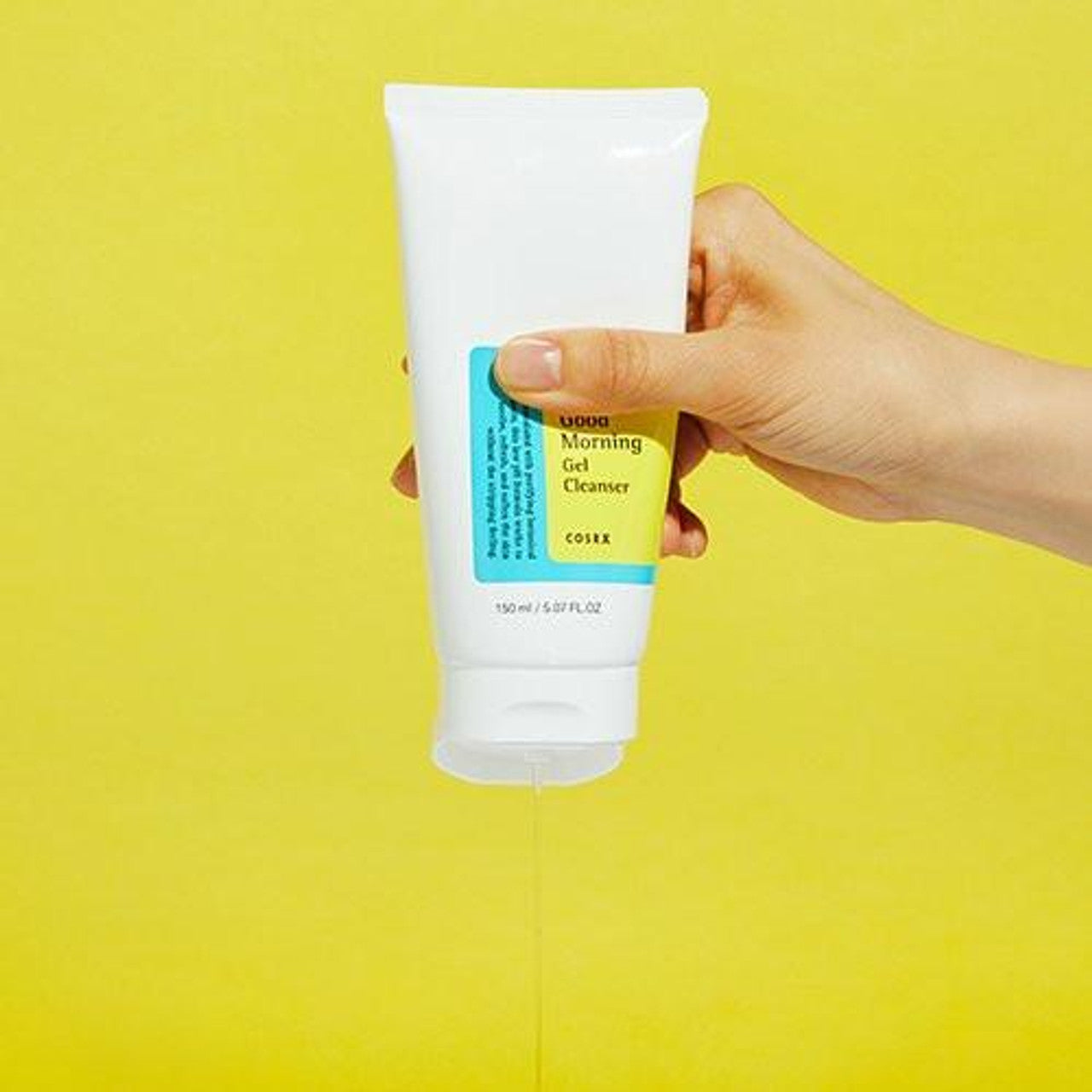 A hand gently squeezes a white tube of COSRX Low PH Good Morning Gel Cleanser 150ml against a yellow backdrop, releasing refreshing gel. This vegan, cruelty-free cleanser with a blue and yellow label promises a rejuvenating start to your day.