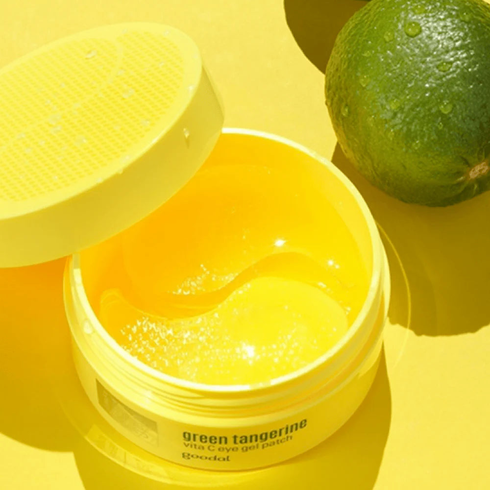 An open jar of Goodal Green Tangerine Vita-C Eye Gel Patch 72g showcases the yellow patches aimed at fighting dark circles. The lid lies beside it, accompanied by a fresh green tangerine on a vibrant yellow background, emphasizing vitamin Cs power.