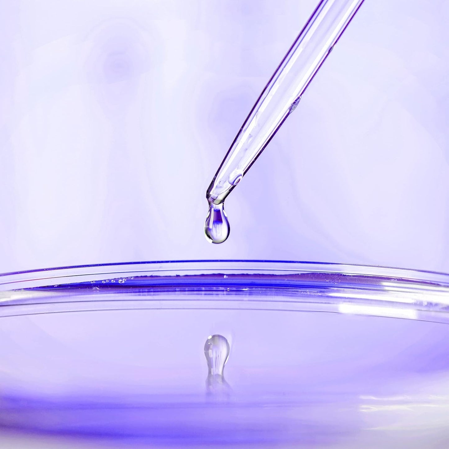 A pipette releases a droplet of liquid into a clear petri dish against a softly lit purple background, creating a scientific and calming atmosphere reminiscent of the soothing effect of OLAYs Cleansing & Renewing Nighttime Body Wash with Vitamin B3 and Retinol reflected in the liquid surface.