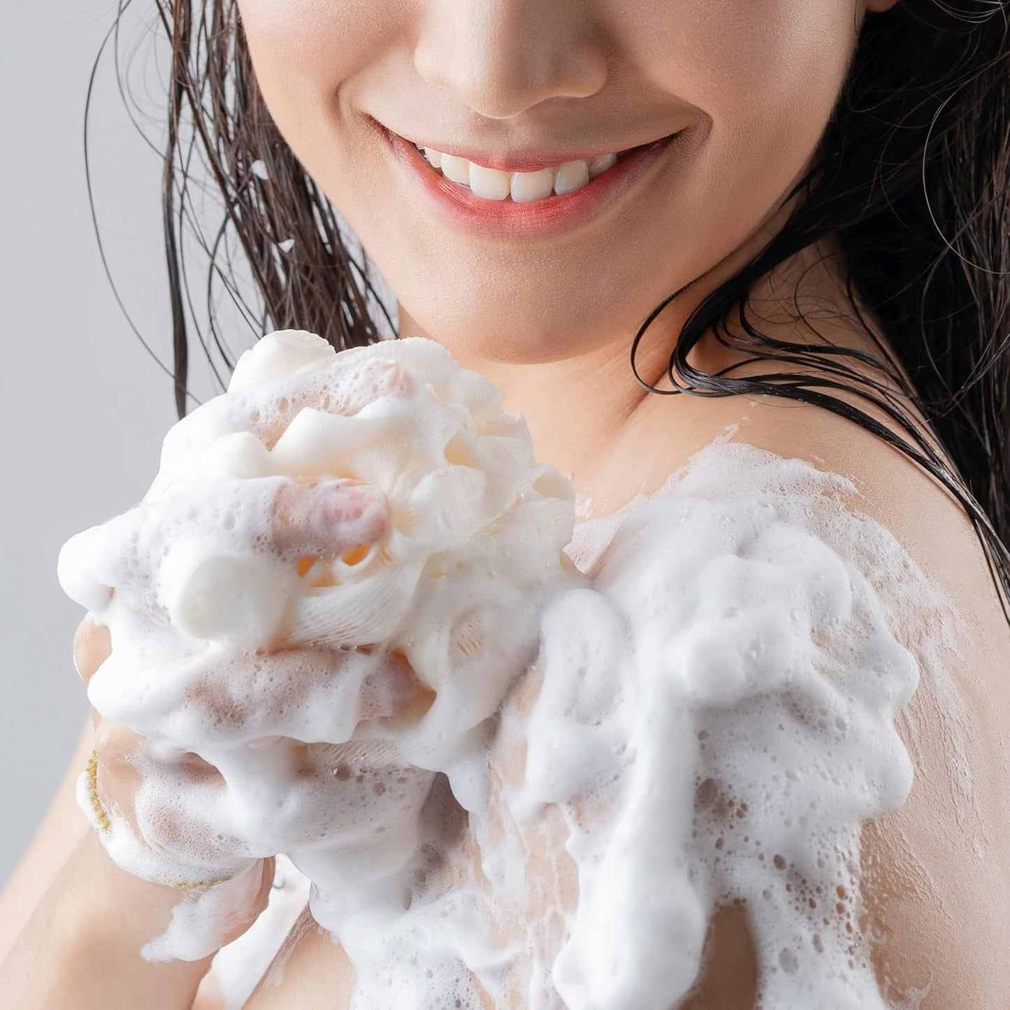 A person with wet hair smiles while applying Olay Cleansing & Renewing Nighttime Body Wash, with Vitamin B3 and Retinol, using a bath sponge. White foamy soap bubbles cover the sponge and part of their skin against a light gray background.