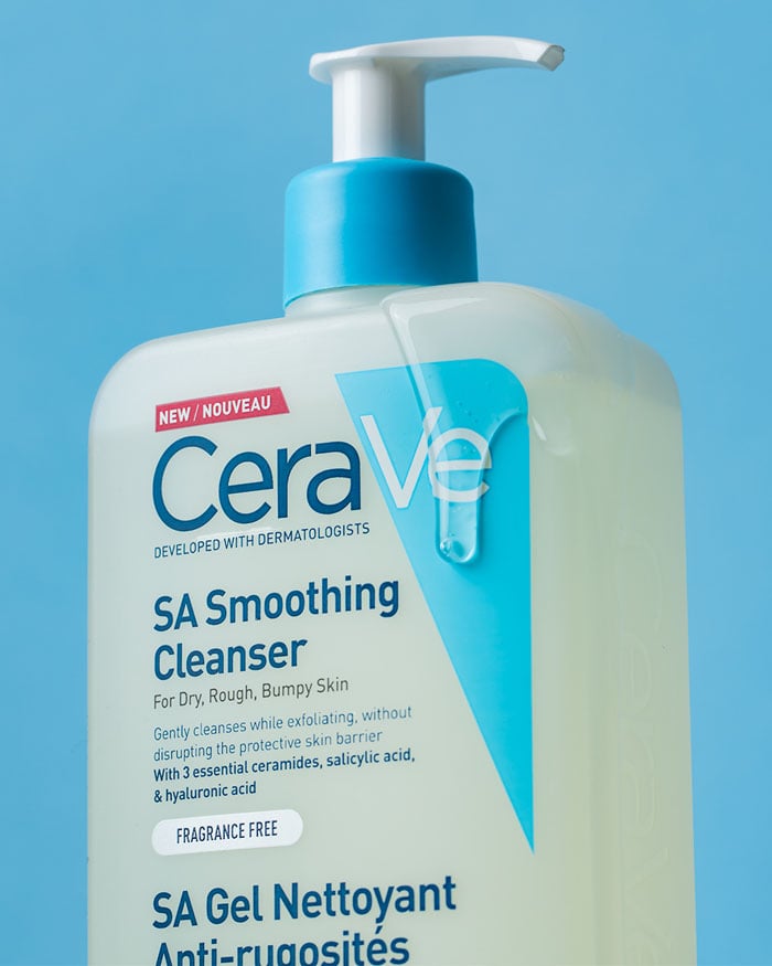 A close-up of a large bottle of CeraVe SA Smoothing Cleanser with Salicylic Acid and Ceramides, featuring a pump dispenser against a blue background. This fragrance-free cleanser is ideal for dry, rough, or bumpy skin.