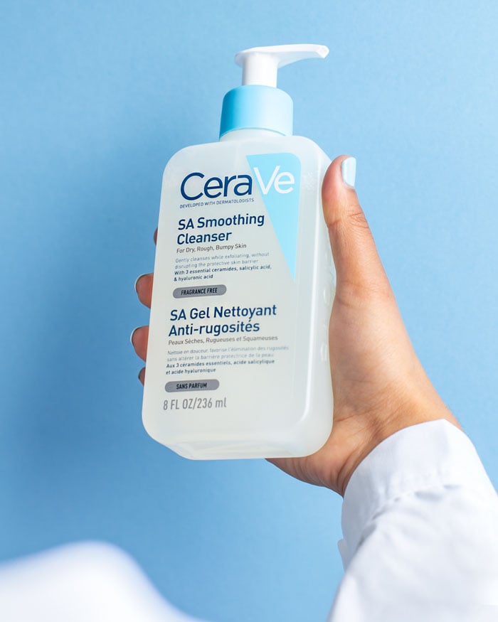 A hand holds a bottle of CeraVe SA Smoothing Cleanser with Salicylic Acid and Ceramides against a light blue background. This fragrance-free cleanser by CeraVe is ideal for rough, bumpy skin and those with keratosis pilaris.