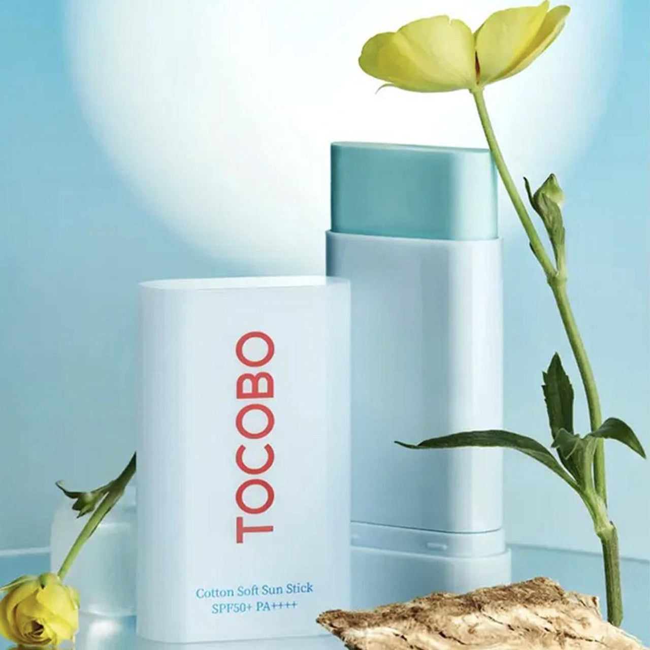 The Tocobo Cotton Soft Sun Stick SPF50+ PA++++ (19g) is elegantly displayed with sleek design alongside a vibrant yellow flower and textured driftwood, set against a soft blue background that enhances its fresh, clean, and vegan appearance.