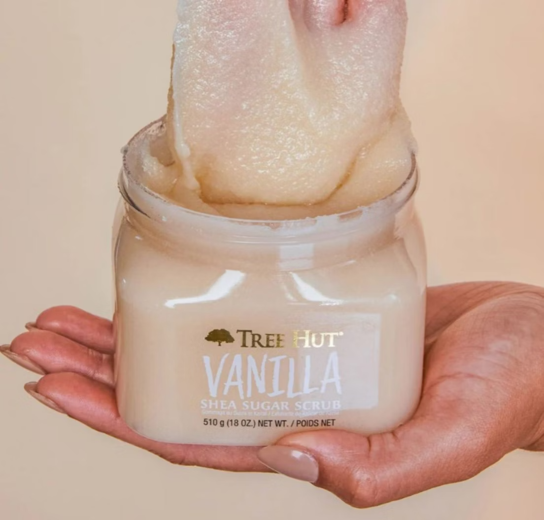 A hand holds an open jar of Tree Hut Shea Sugar Body Scrub - Vanilla 510g, renowned for its rich shea butter blend. Another hand scoops out the off-white, creamy textured exfoliant with a smooth consistency from the container.