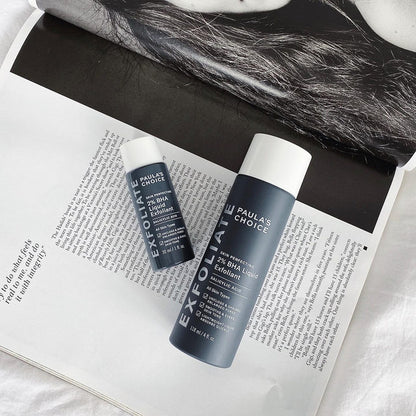 Two sleek bottles of Paulas Choice SKIN PERFECTING BHA Liquid Exfoliant, ideal for blackhead removal, rest on an open magazine with black and white hair imagery. Infused with salicylic acid, this modern product exfoliates dead skin effortlessly.