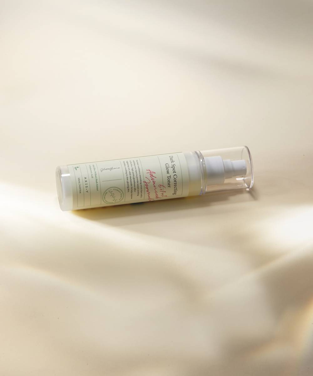 The Axis-Y Dark Spot Correcting Glow Toner, in a clear plastic bottle with a pump, rests on a cream-colored surface. Its minimal label features green and red text. Light and shadow add depth to the background, enhancing its elegant simplicity.