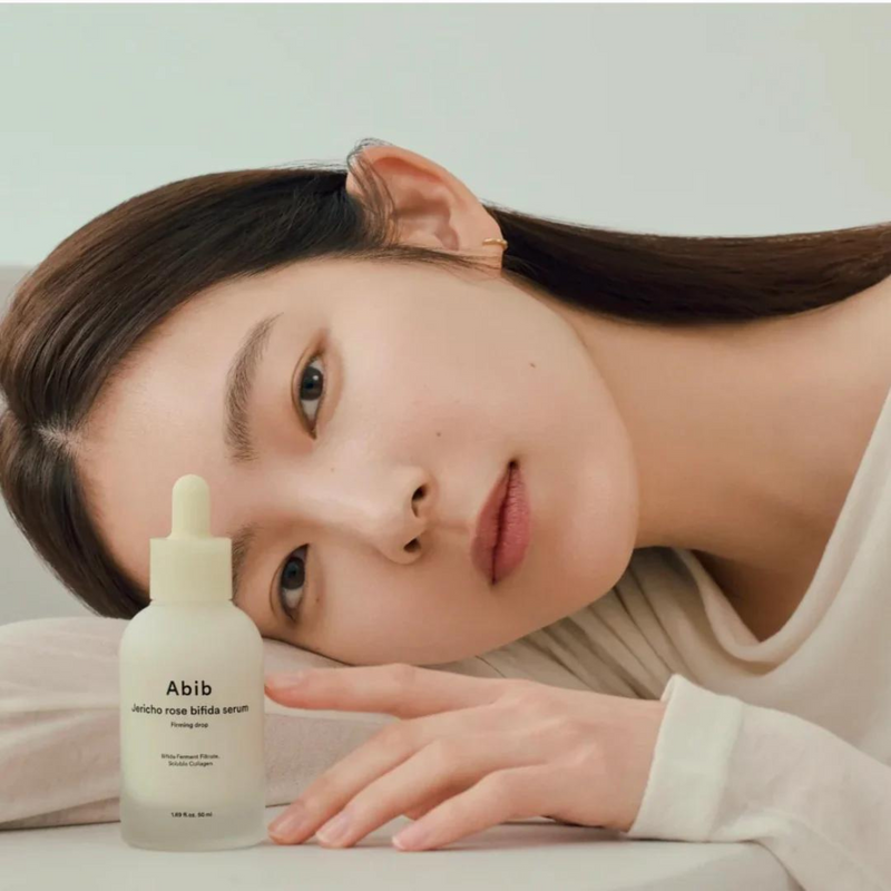 A woman with clear, radiant skin resting her head on a surface, gently touching a bottle of ABIB Jericho Rose Bifida Serum Firming Drop 50ml, highlighting skincare benefits