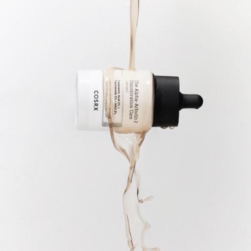 The bottle of COSRX The Alpha-Arbutin 2 Discoloration Care Serum 50ml is suspended against a light background, with a stream of Niacinamide-infused liquid elegantly splashing down, creating a dynamic visual.