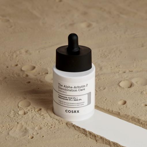 The COSRX The Alpha-Arbutin 2 Discoloration Care Serum 50ml with a dropper cap sits on a textured sandy surface, highlighting ingredients such as niacinamide, mandelic acid, and tranexamic acid for improved skin clarity.